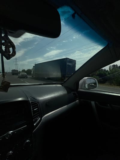 The image is a point-of-view shot from inside a car, looking forward through the windshield. The car appears to be on a highway, with other vehicles visible. A large, dark-colored truck with the license plate "308679AA" is in the right lane. A dark sedan is in the left lane, and another truck is in the distance on the left. The sky is cloudy and blue, with power lines visible. The interior of the car is dark, with the dashboard and part of the steering wheel visible in the foreground. A hanging air freshener or ornament is visible in the upper left corner. The scene suggests travel on a sunny day in a moderately developed area, possibly on the outskirts of Istiqlol, Uzbekistan, given the style of vehicles and infrastructure.