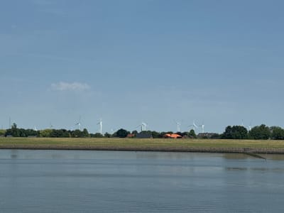The image depicts a scenic view of a canal or waterway in Brunsbüttel, Germany, on a clear, bright day. The foreground is dominated by the calm, rippling surface of the water, reflecting the pale blue sky. A stone embankment separates the water from the land above.

The middle ground features a lush green grassy slope leading up to a line of trees. Dotted among the trees are several houses with terracotta-tiled roofs, suggesting a residential area. Prominently visible across the landscape are numerous wind turbines, their white structures and large blades standing tall against the horizon, indicating the presence of a wind farm and highlighting renewable energy infrastructure. One of the houses has a small red flag flying.

The background is a vast expanse of clear, pale blue sky, with a single wispy cloud visible towards the left. The overall atmosphere is peaceful and serene, characteristic of a calm day in a rural or semi-rural setting with a focus on both nature and modern energy production. The time of day appears to be late morning or early afternoon due to the bright sunlight and clear skies. There is no visible text in the image.