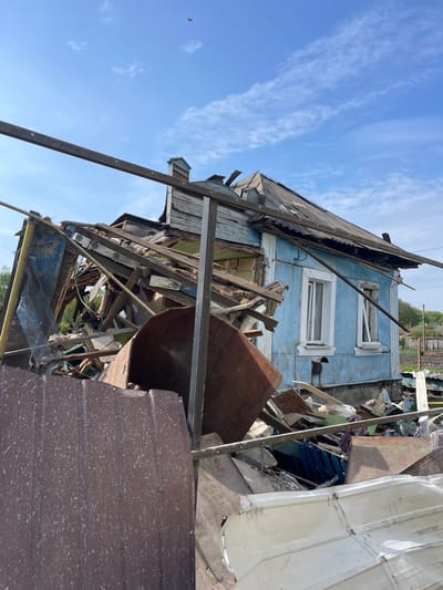 The image shows a partially destroyed building in Bilopillia, Ukraine. The building is a single-story, blue structure with a damaged roof and a broken window. The roof is partially collapsed, and there is debris scattered around the building. The front of the structure is visible, revealing partially boarded up windows and a door. Rubble and debris are piled around the structure. One side of the image includes a large, partially visible metal object. The image is likely taken from the street in front of the building. The image was taken under a blue sky with no visible clouds. The coordinates 51.139, 34.285 place the location in Bilopillia, Ukraine.