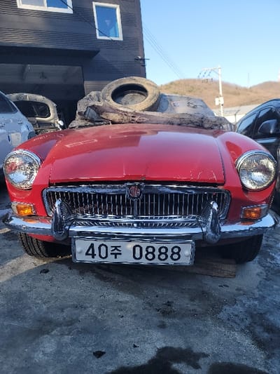A red, vintage MGB roadster faces the camera. It has a chrome grill and bumper with round headlights. A South Korean license plate reads "40 주 0888" vertically. The car appears to be parked outside on concrete. In the background is a dark building, a tire on an object, and hills. The photo has been rotated ninety degrees.
