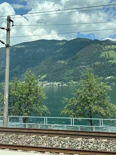 The image depicts a scenic view of Zell am See, Austria, from the perspective of a train. The scene is dominated by a large, dark green, forested mountain that slopes down towards a calm, reflective lake. The water of the lake is a deep teal color, with ripples creating a shimmering effect. Scattered along the lakeshore are buildings that appear to be part of a town or resort, with white facades and red roofs.

In the foreground, on the right side of the image, are train tracks with gravel ballast. A metal railing with a white fence runs parallel to the tracks, separating them from the lake. Two trees with lush green foliage are positioned between the railing and the lake, their branches reaching out towards the water. In the upper left portion of the image, thick, white clouds are visible against a bright blue sky. Several power lines stretch horizontally across the sky, connected to a utility pole that is partially visible on the left.

The image appears to have been taken on a bright, clear day, likely in the summer or early autumn given the greenery. The overall mood is peaceful and serene, capturing the natural beauty of the Austrian Alps and the charming lakeside setting. There are no people visible in the image. There is some faint yellow text on a rectangular object attached to the railing, but it is too blurred to be legible.
