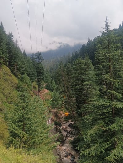 The image depicts a mountainous landscape in Khakhnal, India. A dense forest of tall evergreen trees, primarily conifers, covers the steep slopes of the mountains. A narrow, winding path is visible through the trees, leading down to a rocky stream. The stream appears to be flowing with muddy water. In the distance, the peaks of the mountains are shrouded in clouds. Overhead, several thin, dark cables stretch across the sky, likely part of a cable car system. The overall scene suggests a remote and natural setting, possibly a hiking or recreational area within the region. There are no people visible in the image.