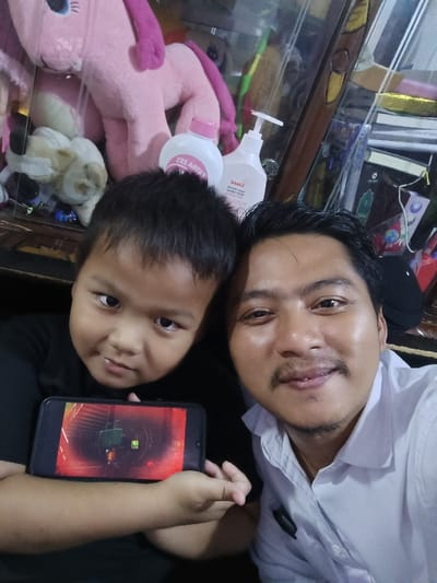 The image displays an adult male and a young male child posing for a selfie in the city of Limbangan Sari, Indonesia. The adult, positioned on the right, has dark hair, a mustache, and light facial hair, wearing a light-colored collared shirt, and exhibits a faint smile directed at the camera. The child, on the left, has dark hair and wears a black shirt, looking towards the camera with a slight smirk. The child holds a black smartphone horizontally, with its screen displaying vivid red and dark-toned graphics, possibly a video game, containing minor green elements. The background, partially obscured by reflections and a transparent barrier, features a large pink plush toy, a smaller light-colored plush toy, two white bottles (one with an inverted "EXTRA 2X" label and a pink cap, the other a pump bottle with "liora" visible), a stack of books, and a dark piece of headwear.