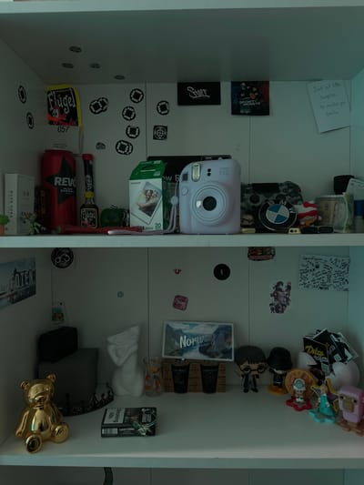 The image depicts a shelf filled with various items, suggesting a personal collection or a curated display. The scene is brightly lit, with the shelf itself appearing to be part of a larger unit, possibly a locker or a display case, given the white, smooth surfaces and vertical dividers.
The subjects are predominantly objects, with a few small figures and no clear depiction of people. On the left side of the shelf, a light pink Instax Mini 12 camera is prominently displayed, with a pack of Fujifilm Instax Mini film next to it. Above the camera, a small square artwork or sticker with the text "SPORTY MONSTR" is visible. To the left of this, a handwritten note reads "Just a little surprise to make you smile". Further down on the left, a red can with the "REVO" logo is present, along with a small bottle of "Flügel". There are also several small circular stickers with a checkered pattern scattered across the shelf.
On the right side of the shelf, a collection of small figurines is arranged, including what appears to be characters from popular culture, such as Harry Potter. There are also some stacked items resembling boxes or packaging, one with the text "Norway". A pack of "Lucky Strike" cigarettes is also visible, with the warning "Smoking kills". A golden teddy bear figurine lies on its side at the bottom right.
The overall impression is one of a personal, eclectic collection of memorabilia, trinkets, and everyday items. The context of Roskilde, Denmark, does not provide specific visual cues within the image itself, but it grounds the location of this scene. There are no clear indicators of time of day or weather, and the emotions conveyed are neutral, stemming from the arrangement of objects.