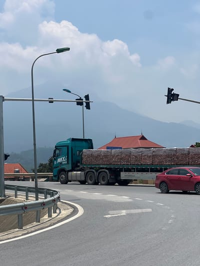 In Tu Vũ, Vietnam, a teal-colored semi-trailer truck, branded "MAX" with license plate "199-042.33", transports a large load of stacked, wrapped wooden planks. The truck is driving on a paved road that curves to the right. A red sedan is visible to the right of the truck, also on the road. Overhead, traffic lights and streetlights are mounted on poles. In the background, a building with a red roof is situated against a backdrop of hazy, layered mountains under a sky with scattered clouds. A metal guardrail lines the left side of the road.