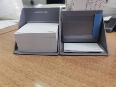The image displays two gray holders on a wooden desk. The holder on the left contains a stack of white paper, possibly sticky notes, with "IREKS" printed on the top sheet. The words "www.ireks.com" are visible on the inside wall of this holder. The holder on the right contains a smaller stack of white paper and a blue sticky tab attached to its inner wall. A piece of paper with handwritten text in what appears to be Cyrillic script and the number "080" is partially visible in the upper right portion of this holder. The desk surface is made of light brown wood with visible grain. The background shows a blurred white object and a dark electronic device. There are no people visible in the image, and no specific location in Dobrodan, Bulgaria can be identified.