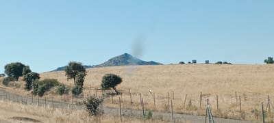 This image captures a dry, grassy hillside under a clear blue sky. A rocky peak dominates the background, with a plume of dark smoke rising from its summit, suggesting a fire. Scattered trees with dense green foliage are present on the hillside, along with a fence line in the foreground. Several dark, rectangular structures, possibly water tanks or small buildings, are visible on the distant horizon. The ground is covered in dry, yellowish grass, indicating a dry climate. There are no people visible in the image. This scene appears to be in a rural, mountainous area. While the image shows a landscape, there is no specific information to pinpoint its exact location within the city of Santa Cruz de la Sierra, Spain.
