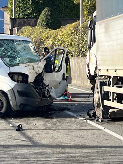 The image captures the aftermath of a multi-vehicle collision on an asphalt road in Griante, Italy. A white van on the left displays severe frontal impact damage, including a completely shattered windshield, a crumpled hood, and a demolished front bumper exposing internal engine components. The driver's side door of the van is open. Adjacent to it on the right is a large white truck, partially visible, which also exhibits damage to its front wheel well area.
One individual, identified as emergency personnel, is visible near the open door of the white van, wearing a dark helmet and a blue uniform with reflective elements. Another person with a yellow-striped uniform is partially obscured behind them. Scattered on the road surface between and around the vehicles are various debris, including broken vehicle parts, glass fragments, and dark fluid stains. A blue and red hydraulic rescue tool is positioned on the ground near the van's open door. A single black boot or shoe lies on the asphalt in the immediate foreground. The environment includes a concrete retaining wall in the background, backed by dense green bushes and trees, under clear daylight conditions.