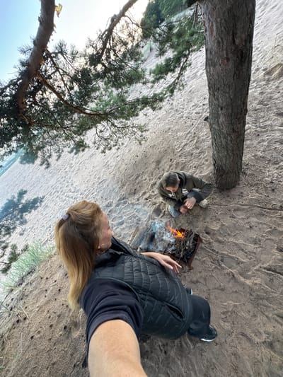 In Fort-Krasnaya Gorka, Russia, on a sandy beach near a pine tree, two individuals are present. One person, in the foreground and on the left, is a woman with blonde hair tied back, wearing a black puffer vest over a black sweatshirt and black pants. She is looking towards a small bonfire in a metal fire pit located in front of her. The other individual, in the middle ground and on the right, is crouching near the bonfire, wearing a green hooded jacket and grey pants, and appears to be tending to the fire. The sandy ground is textured with footprints and scattered debris, and a portion of the sea and a distant treeline are visible in the background. The scene is captured from a slightly elevated, wide-angle perspective.