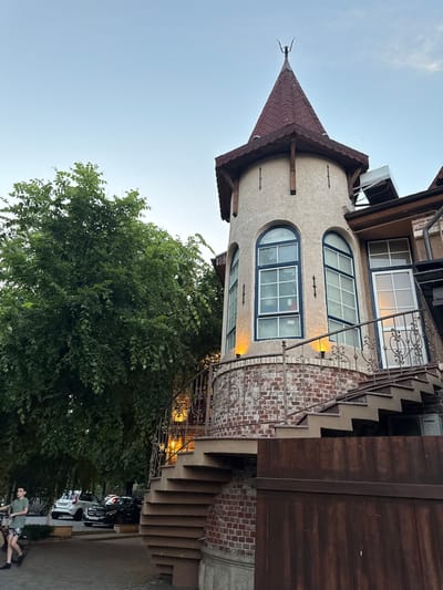 This media file features a tall, multi-story building with a distinctive turret and a conical, shingled roof. The building has arched windows with blue frames and a decorative wrought-iron railing. Adjacent to the turret, there are brick walls and an outdoor staircase leading upwards. A large, leafy green tree partially obscures the lower section of the building. In the background, parked cars are visible, and a person is seated on a bicycle in the foreground.

The scene appears to be set in the evening, indicated by the warm glow of lights emanating from within the building and the soft, muted sky. The weather seems calm and clear. The location context is Oktyabr'skoe, Russia. The overall mood is relaxed and potentially hints at a residential or commercial setting with an inviting ambiance. There is some visible text on a white appliance mounted on the building, which appears to say "GoldStar".