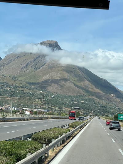 The image depicts a multi-lane highway on a partly cloudy day, with a large, rugged mountain dominating the background. The scene is consistent with the landscape near Termini Imerese, Italy, with the prominent mountain likely being Monte San Calogero.

In the foreground, an asphalt highway stretches into the distance, featuring at least two visible lanes of travel marked by white dashed lines. A vegetated median strip, containing low green shrubs and secured by metal guardrails, separates the direction of travel from the opposing lanes. On the visible side of the highway, a dark grey SUV is positioned in the right lane, with a red car following behind it. On the opposite side of the median, a bright red tour bus is prominent in the left lane, with several other vehicles visible further down the road. A green road sign displaying "100" is visible on the right shoulder of the highway, suggesting a speed limit or distance marker.

The middle ground transitions from the highway infrastructure to the lower slopes of the mountain. These slopes are extensively cultivated with dense olive groves, characterized by numerous small, rounded green trees. Scattered among the groves are various small buildings and residential structures. Streetlights with modern fixtures line sections of the highway.

The background is dominated by a massive, steep mountain. Its upper sections feature exposed grey rock formations, while lower parts are covered in sparse green vegetation. Patches of white cumulus clouds are draped across the mountain's mid-section and summit, partially obscuring the highest peaks. The sky above the mountain is a clear, light blue. No individuals are directly visible in the immediate foreground or on the highway.