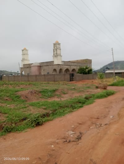The image depicts a mosque in Ibo, Nigeria, under an overcast sky. The mosque features a light-colored facade with arched windows and two prominent minarets topped with golden domes. A concrete wall surrounds the mosque, and a black metal gate is visible. In the foreground, there is an undeveloped area with patchy green grass and reddish-brown dirt, suggesting a rural or developing urban setting. A dirt path leads into the scene. Power lines are visible in the sky above. The timestamp "2026/06/13 09:09" is overlaid in the lower left corner of the image.