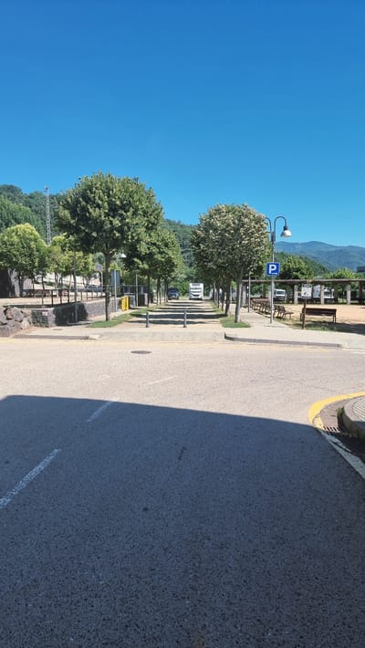 The image shows a street scene in Castellfollit de la Roca, Spain. The perspective is from the foreground, looking down a tree-lined avenue that leads towards a body of water and mountains in the distance. The street is paved with asphalt and marked with white lines. On the left side of the avenue, there is a stone wall and some park benches. On the right side, there are more park benches and a parking sign. Several vehicles, including a caravan and a black SUV, are parked along the avenue. The sky is clear and blue. The overall impression is one of a peaceful, sunny day in a scenic location.