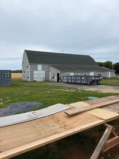 This image shows a construction site with a large, gray building in the background and wooden materials in the foreground. The building appears to be a barn or a similar agricultural structure, with a dark green roof and light gray siding. There are several windows and doors on the building, some of which are boarded up. A large, dark gray dumpster is parked next to the building, with "SUNRISE" written on its side in white letters, along with a phone number.
In the foreground, there are various construction materials, including large sheets of plywood and wooden beams. Some of the wood appears to be freshly cut, while other pieces show signs of wear. There is also a pile of gravel and a section of what looks like a concrete curb or foundation. The ground is covered in grass and dirt. The scene appears to be an outdoor construction project.
The sky is overcast, suggesting it is either early morning or late afternoon, or a cloudy day. There are no people visible in the image. The overall atmosphere is one of ongoing work or preparation for construction. The location is identified as Mattituck, United States of America.