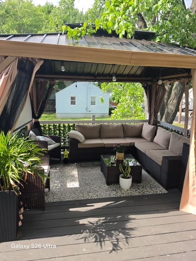 The image depicts a backyard gazebo on a wooden deck, situated in West Hartford, Connecticut, USA. The gazebo features a brown metal roof and a tan canopy with mesh sides. Inside, a large sectional sofa with beige cushions is arranged around a dark wicker coffee table. A potted plant sits on the coffee table. To the left, a wicker chair with patterned cushions is visible, with a bright yellow electric leaf blower on the deck in front of it. A large potted plant with green fronds stands in the foreground on the left. Behind the gazebo, a light blue house with white trim and windows is visible, surrounded by green grass and trees. The deck shows signs of wear, with shadows cast by the gazebo's roof and surrounding trees. The overall scene suggests a residential outdoor living space.