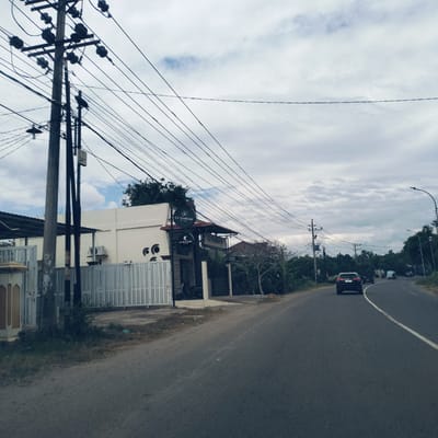 This image captures a street scene in Gampongrayadagang, Indonesia, featuring a road, buildings, and an extensive network of utility poles and overhead power lines. The asphalt road curves gently to the right, with a solid white line visible on its surface.

On the left side of the road, a cream-colored commercial building is set back behind a grey slatted fence. The building has an air conditioning unit mounted on its exterior. Several dark circular signs are affixed to the front, displaying white text: one prominent sign reads "Cr Fashion IN STORE," and another smaller one below it says "Cr Kosmetik." A motorcycle is parked within the premises, visible through the entrance gate.

The sky above is overcast with scattered clouds, suggesting daytime conditions. A dense array of utility poles and crisscrossing power lines fills the upper left portion of the frame. Two birds are distinctly visible, perched on different parts of these utility poles.

Further down the road on the right, several vehicles are present, including a black SUV in the foreground followed by other cars and a van. The roadside is lined with trees and lush green foliage, with additional streetlights visible in the distance. The overall scene depicts a functional, everyday environment in a semi-urban or suburban setting.