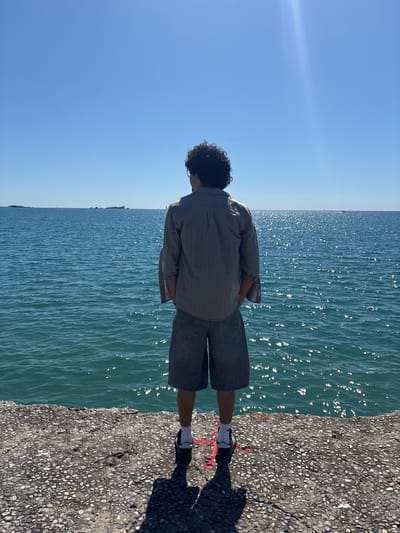 A person stands on a concrete pier or walkway bordering a body of water, likely the Mediterranean Sea, under a clear blue sky. The person is facing the water, with their back to the viewer. They are wearing a striped button-down shirt, dark denim shorts, white socks, and dark shoes. The concrete pier has a pebbled surface. The water reflects the sunlight, creating a shimmering effect. The location is likely in Fréjus, France, given the typical coastline and light.