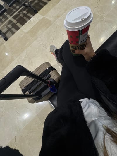 An individual is depicted from a high-angle perspective, seated in what appears to be a public waiting area, such as an airport or bus terminal lounge. The precise location within San Miguel Totoltepec, Mexico, cannot be determined from the generic architectural features. The person wears black leggings, light-colored athletic shoes with multi-colored accents, and a black fuzzy jacket over a white top. Their right hand holds a red, holiday-themed Starbucks cup with a white lid and a brown cardboard sleeve. A ring is present on their right ring finger, and dark nail polish is visible. Adjacent to the person, a light brown hard-shell rolling suitcase with a black extended telescopic handle rests on the floor; a small blue and red fluffy keychain is attached to the handle. The floor is composed of light beige or cream-colored polished stone tiles with subtle veining. Partial views of dark, utilitarian seating are visible in the background, casting distinct shadows on the tiled floor, indicating artificial lighting. No other individuals or specific interactions are visible.