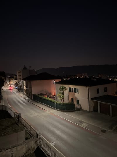 The image captures a nighttime street scene in Balerna, Switzerland. A gently curving road, illuminated by streetlights, dominates the foreground and midground. The road surface appears wet, reflecting the artificial light. Red lines mark the edges of traffic lanes, and several circular manhole covers are visible. A dark metal fence runs along the left side of the road, separating it from a slightly raised, dark grassy area.

On the right side of the road, a light-colored building with dark-shuttered windows stands prominently. A modern streetlight illuminates the area in front of this building. A well-maintained hedge borders the property. To the right of this building, a structure with a dark roof and what appear to be garage doors is visible. Further down the street, several vehicles are parked.

In the background, other buildings and streetlights indicate a residential or commercial area. Beyond the built environment, the silhouette of a dark, tree-covered hill or mountain range is visible against the night sky. The sky itself is dark, with a faint gradient of light towards the horizon, suggesting a clear night. Two cars are visible on the road, one with its red taillights illuminated, heading away from the viewer. One car is red, and appears to be moving away from the viewer, with its taillights illuminated.