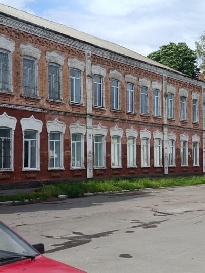The image captures a section of a two-story brick building with a light-colored roof, located on the street in Khorol, Ukraine. The building's facade features rows of windows, some with decorative white trim. The lower story windows have metal bars. Green grass grows along the base of the building, bordering a paved road. In the foreground, the hood and windshield of a red car are visible. The sky above is partly cloudy.