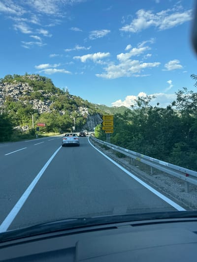 A slightly rotated medium shot captured from inside a car moving along a highway in Donji Ulići, Montenegro, presents a clear, bright day scene. A guardrail runs along the right edge of the road. A white vehicle is in the middle lane, preceded by a black vehicle further ahead. Road signs indicate directions to Mikulici, Kostajeri, Bokovo, Dinovici, and Markov Do. A hillside covered with trees and rocky outcrops stretches along the left side of the road. The sky above is bright blue with scattered white clouds.