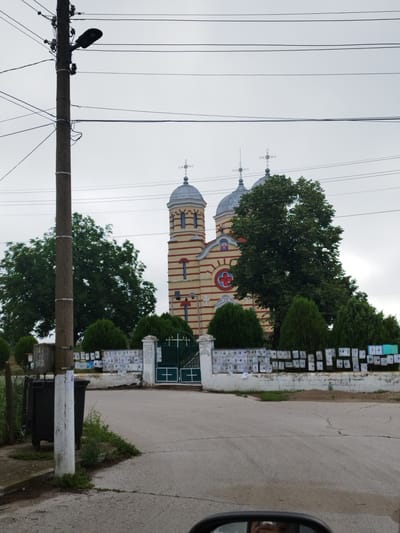 The image shows a street scene in Slanotran, Bulgaria, with a church in the background. A concrete utility pole with a street lamp and overhead wires is prominent in the foreground on the left. To the right of the pole, a dark gray trash bin is visible. A white concrete wall with numerous posters attached lines the perimeter of the church grounds, which also feature a green metal gate. Behind the wall, manicured evergreen bushes and large trees are present. The church itself has a distinctive yellow and red striped facade, two domes topped with crosses, and arched windows. The sky is overcast. The image appears to be taken from a vehicle, as suggested by the reflection in what seems to be a rearview mirror at the bottom of the frame.