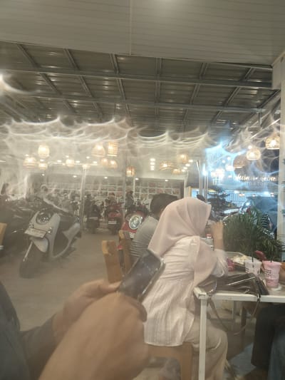 The image depicts an indoor-outdoor seating area at what appears to be a cafe or restaurant. The scene is illuminated by warm, hanging rattan lamps, creating a cozy atmosphere. Through a glass partition, several motorcycles and scooters are visible parked, suggesting a mode of transportation common in the location or the establishment's parking setup.
In the foreground, a person's hands are holding a smartphone, indicating someone is engaged with their device. To the right, a woman wearing a light pink hijab and a matching textured dress is seated at a white table. Her attention seems to be directed away from the camera. On the table, there are plastic cups with pink beverages, a bag, and some snacks or food items, suggesting people are dining or enjoying drinks.
Further into the background, more patrons are seated, and other motorcycles are parked. The overall impression is a casual gathering spot, possibly a cafe that caters to customers arriving by motorcycle. The lighting suggests it could be evening or a dimly lit indoor space. The presence of hanging lamps and the style of the cafe suggest a modern but relaxed ambiance.
Visible text on a sign in the background, partially obscured by reflections and the haze, appears to read "Wulan Kebal" followed by "RO & COFFEE". The time of day is likely evening or night due to the artificial lighting and the dimness of the natural light. The weather is not discernible from the image. The setting appears to be in Blangbitra, Indonesia, as per the provided context.