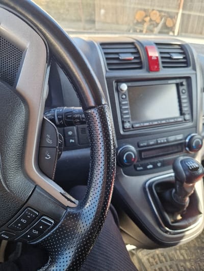 The image depicts the interior of a vehicle, presumed to be located in Kasak, Bulgaria, viewed from the driver's perspective. The foreground is dominated by a black, perforated leather steering wheel with silver-grey plastic spokes and integrated multi-function controls on the left. A driver's leg, clad in dark, ribbed fabric, is visible beneath the steering column in the lower left quadrant. The dashboard features a central console with two air vents, a prominent red hazard light button, an infotainment unit with a dark screen and control buttons, and manual climate controls. A six-speed manual gear shifter, encased in a black leather boot, is positioned to the lower right of the center console. Through the windshield, a blurry background reveals an outdoor scene with a wooden fence and a stack of cut logs, illuminated by natural daylight.