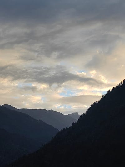 The image depicts a mountainous landscape at dusk, likely in the vicinity of Old Manali, House of Israel, India. The foreground is dominated by the dark silhouettes of several mountain ranges, layered one behind another. The closest mountain range on the right is covered with trees, whose silhouettes are clearly visible against the lighter sky. The sky itself is a gradient of blues and grays, with streaks of sunlight breaking through the clouds, illuminating parts of the sky with a soft, warm glow. The overall mood is serene and dramatic, characteristic of a mountain sunset or sunrise. No people or specific structures are visible, focusing solely on the natural environment.