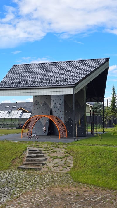 This is a vertical, outdoor, eye-level shot of a modern climbing wall structure, located in Kamenka, Russia. The climbing wall is an angular, grey structure with geometric patterns, featuring numerous colorful holds for climbing. To the left of the wall, an orange, semi-circular net structure encloses a child's bicycle and two hanging ropes. The climbing wall is situated under a dark, grey, tiled roof with a steep pitch, supported by a white awning. In the background, other buildings with similar roofs and a greenhouse are visible, along with mature trees and a fence. The foreground shows a sloped lawn with irregularly shaped stone steps leading up to a paved area. The sky is blue with scattered white clouds. The scene appears to be a public recreational area.