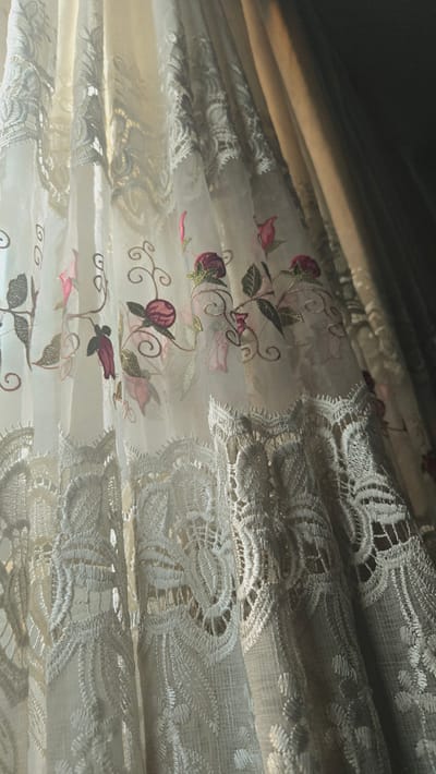 The image displays a close-up view of an embroidered sheer curtain with a floral pattern. The embroidery features delicate roses in shades of pink and burgundy, along with green leaves and brown tendrils. The lower portion of the curtain is adorned with intricate white lace. The curtain is hung in a room, with a darker, heavier fabric visible behind it, suggesting it is layered. Sunlight streams through the sheer fabric, creating soft shadows and highlighting the textures of the embroidery and lace. The overall impression is one of elegance and natural light. There are no people or indications of the specific location within Kaka, Turkmenistan, in the image.