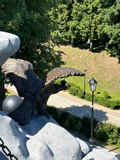 The image shows a bronze eagle sculpture perched on a granite base in Vereya, Russia. The eagle has its wings spread and is facing left, with its head turned slightly downwards. A spherical object is located to the left of the eagle, partially obscured by its wing. In the background, there is a path lined with hedges and trees. Two black lampposts with white lights are visible, one closer to the foreground and another further back. A security camera is mounted on the pole of the closer lamppost. The ground is covered with dry grass and some paved areas. The scene is illuminated by natural sunlight, casting shadows.