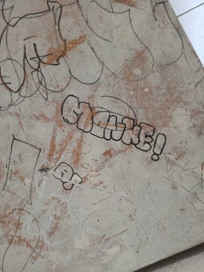 The image depicts a close-up, angled view of a worn, light-colored wooden or composite surface, identified as being in Kawungluwuk, Indonesia. The surface exhibits extensive black and white marker graffiti, scratches, and areas where an underlying reddish-brown material is visible. The primary subject is a black outline bubble letter tag spelling "CHANE!" followed by an exclamation mark, positioned diagonally across the mid-section. Below this, a smaller black outline tag resembling "QQ" or "QO" is present. Numerous other irregular black line scribbles and indistinct outlines cover the surface. In the lower-left quadrant, a roughly drawn black rectangle contains faint illegible text, including the discernible word "Bear". White marker lines are also visible, some overlaying black markings. No individuals are present in the scene; the actions observed are the application of graffiti to the surface. The environment suggests an urban or semi-urban setting, likely a public or semi-public space where such markings would occur.