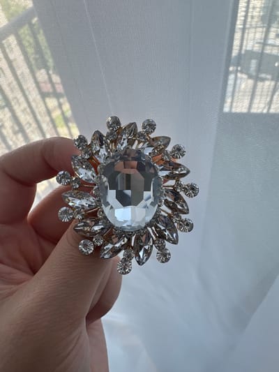 A hand holds a sparkling brooch with a large central gem, surrounded by smaller faceted stones, against a sheer white curtain that allows light and the indistinct shapes of a window with blinds to show through. The brooch has a floral or sunburst design, with gold-toned metalwork. The hand is relaxed and fingers are slightly curled. The background is a soft, out-of-focus blur of light and shadow. The scene appears to be indoors, possibly in a residential setting.