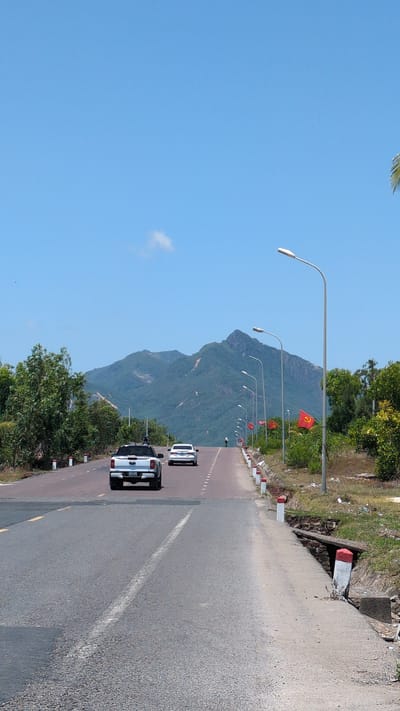 This image depicts a sunny, outdoor scene on an asphalt road in Tuy Phong 4, Vietnam, leading towards a mountainous backdrop. The road exhibits a slight incline and features two visible lanes in the foreground, separated by a solid white line and a dashed yellow line on the left. A concrete and dirt shoulder with white and red painted concrete markers runs along the right edge of the road, revealing a partially collapsed concrete culvert.
In the foreground, a white Ford Ranger pickup truck with a visible license plate is moving away from the viewer in the right lane, with a black object mounted on its roof. Further ahead, a white sedan is present in the left lane, also traveling away. In the distant center of the road, a lone individual on a dark motorbike is visible.
Along the right side of the road, a series of tall, grey metallic streetlights with white fixtures extends into the distance. Between these streetlights and dense green vegetation (trees and bushes), several red flags bearing a yellow hammer and sickle emblem are affixed to poles. Similar vegetation lines the left side of the road. The background is dominated by large, verdant mountains beneath a clear, bright blue sky containing a single small white cloud.