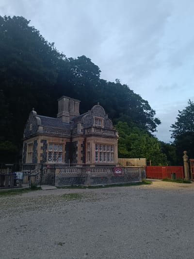 A wide shot of a historic building, likely a lodge or gatehouse, with distinctive stonework and ornate windows, stands against a backdrop of dense green trees and a cloudy sky. The building's facade features alternating bands of dark and light stonework, with numerous multi-paned windows. A decorative balustrade runs along the front of the building. To the right, a bright orange wooden fence encloses an area with some greenery. In the foreground, a gravelly surface extends towards the building, with a black metal fence and gate providing access. A sign with two swan silhouettes is visible on the balustrade. The scene appears to be in a natural setting, possibly near a park or woodland area. The lighting suggests it is either early morning or late evening. No people are visible in the image. The location is identified as Arun, United Kingdom, though specific city or landmark details are not provided by the image alone.