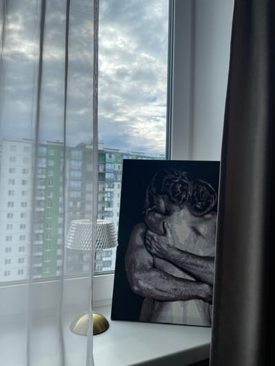 The image shows a painting of two embracing figures, rendered in grayscale, placed on a windowsill. To the left of the painting is a small, modern lamp with a textured diamond-patterned shade and a brass base. The windowsill overlooks a city landscape with apartment buildings, some of which are brightly colored green and white, suggesting a residential area. The sky is cloudy and overcast. Sheer white curtains hang to the left, and a dark gray curtain is visible to the right, partially obscuring the view. The overall scene is indoors, on a windowsill, with an urban background. The painting is the central subject, capturing a moment of intimacy between the two figures.