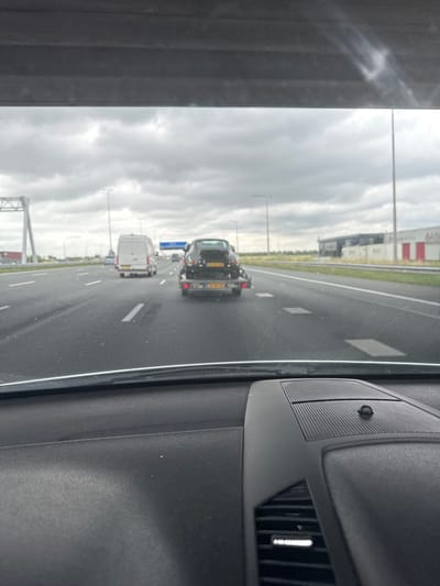 The image is taken from inside a vehicle, looking out at a highway scene. The perspective is from the passenger seat, with the dashboard of the car visible on the right side of the frame. The dashboard includes a speaker grille, an air vent, and a small black circular object. The highway is wet, suggesting it has recently rained or is raining.

The highway has multiple lanes, with several vehicles on the road. The most prominent vehicles are a dark-colored truck or large SUV in the middle lane and a white van in the lane to its left. Both vehicles appear to be moving forward. In the distance, more vehicles are visible, as well as tall streetlights and some industrial-looking buildings on the left side of the road. The sky is overcast and cloudy, indicating a gray or possibly rainy day. The overall lighting suggests it is daytime.

There are no people clearly visible in the image. The scene appears to be a typical highway commute or travel on a cloudy day. Some text is visible on the license plate of the leading vehicle, but it is not legible. The location context provided is Hoofddorp, Netherlands.