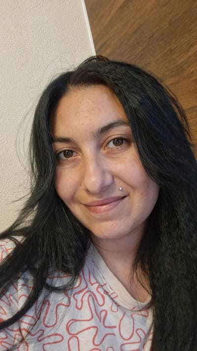 This is a portrait-style, medium shot image of a woman with long, dark, wavy hair. She is looking towards the camera and smiling slightly. She has brown eyes, a light complexion, and a small silver stud piercing on her left cheekbone. She is wearing a light-colored shirt with a repeating pattern of red, abstract, wavy lines. The background is split between a textured, light-colored wall on the left and a wooden surface on the right, suggesting an indoor setting, possibly a room in a residential building. The image does not contain any information to place it specifically within Štětí, Czechia.