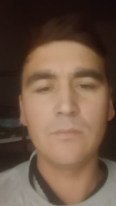 The image presents a close-up, head-and-shoulders portrait of an adult male. He has short, dark brown hair and dark, slightly downcast eyes under prominent eyebrows. His nose is straight, and his lips are closed in a neutral expression. He wears a light-colored, possibly grey, garment with a dark collar or trim visible at the neck. The background is dark, blurred, and indistinct, suggesting an interior setting with faint shapes of furniture or objects. Lighting is dim and appears to emanate from the front. The image contains no discernible landmarks or features to visually confirm the stated location of Karasu, Uzbekistan.