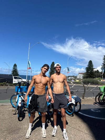 The scene depicts two shirtless adult males standing side-by-side on a paved walkway at Bondi Beach, Australia, under a clear blue sky with sparse clouds. Both individuals wear dark athletic shorts, white socks, and light-colored athletic shoes. The individual on the left has dark hair, wears a black backward baseball cap, a thin necklace, and holds a black garment over his left forearm. The individual on the right has lighter hair, wears a white backward baseball cap, and has white wired earbuds. Immediately behind them is a row of blue and white public share bicycles, with a partial green bicycle visible on the far right. Further back, a street runs parallel to the walkway, separated by a decorative metal barrier. Blue and white flags are attached to poles along the street, and overhead streetlights are visible. A white utility vehicle is parked on the street to the left. In the background, evergreen trees line the coast, the Pacific Ocean is visible, and white residential buildings are present further afield.