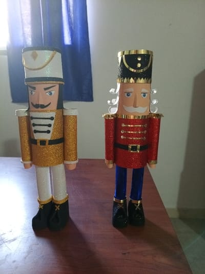 The image displays two decorative, glitter-covered soldier figures, resembling Nutcrackers, standing upright on a polished wooden surface. The surface exhibits a reddish-brown hue and visible wood grain, with shadows cast by the figures indicating a light source from the left.
The figure on the left wears a gold glitter jacket with black and white detailing on the chest, white glitter trousers, and black boots accented with gold bands. Its head features a light skin tone, blue eyes, black eyebrows, a black mustache, and dark hair under a white glitter hat with a black band and gold accents.
The figure on the right wears a red glitter jacket with gold detailing on the chest, blue glitter trousers, and black boots with gold bands. Its head also has a light skin tone, blue eyes, a white mustache, and white curly hair visible below a black glitter hat adorned with gold details.
The background consists of a light-colored wall on the right side and a window with a dark blue curtain on the left, allowing natural light into the indoor setting. No specific identifying features of a city or an exact location within Cumana, Venezuela, are discernible from the visual information provided.