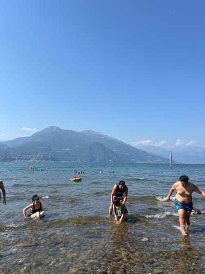 This image captures a daytime scene at the shoreline of Lake Como, near Griante, Italy. The foreground features clear, shallow lake water with a visible rocky bed.
Five individuals are prominent in the foreground. On the far left, a partial view of a person entering the water is visible. To their right, a woman in a dark swimsuit is seated in the shallow water. In the center, a man in a dark tank top stands in the water, appearing to assist a second woman who is kneeling in front of him; she wears a light-colored swimsuit. On the far right, a shirtless man in patterned blue swim trunks walks through the shallow water, generating small splashes.
Further out in the midground, the lake water extends, with several other individuals visible, swimming or wading. A red and green inflatable ring floats in the water to the left of center. A tall, white and dark striped pole stands vertically in the water on the far right.
The background is dominated by a range of large, steep mountains, heavily vegetated, rising directly from the opposite side of the lake. Settlements or towns are visible along the base of these mountains. The sky above is clear and uniformly bright blue, with a small, distant white object visible in the upper right quadrant. The scene is brightly illuminated by direct sunlight, indicating clear weather.