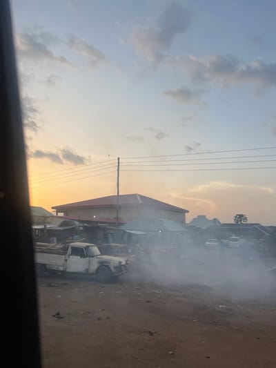 This image depicts a dusty, unpaved street scene at either sunrise or sunset in Toro, Nigeria, characterized by an orange-yellow glow near the horizon blending into a light blue sky with scattered clouds overhead. A dark, out-of-focus vertical object partially obscures the left foreground.
In the mid-ground, an older model white pickup truck, appearing worn and somewhat damaged, is parked facing right, loaded with planks of wood or similar construction material in its bed. A person in a light-colored garment is partially visible near the truck's bed, suggesting activity such as loading or unloading. Behind the truck and extending to the right are various single-story and two-story structures, some with red or corrugated metal roofs, indicative of local shops or residences. A utility pole with multiple overhead power lines stretches horizontally across the upper mid-ground, and a small dark bird is perched on one of these lines against the illuminated sky.
To the right side of the frame, a dense cloud of white smoke or haze emanates from ground level, obscuring the lower portions of two lighter-colored vehicles (possibly cars or small vans) parked further down the street. Additional structures with dark roofs are partially visible through the haze in the background. The ground is consistently reddish-brown dirt with scattered debris. A solitary tree is discernible on the distant right horizon.