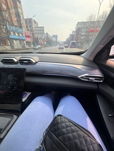 The image presents an interior perspective from a vehicle in Huanggu District, China, looking forward onto an urban street. In the foreground, the legs of an individual are visible, clad in light blue trousers and dark sneakers, with a black quilted handbag featuring a silver-colored strap resting on their lap. The vehicle's dashboard is prominent, showcasing dual air vents with silver accents, a patterned trim on the passenger side, and a large vertical touchscreen display showing the time as 13:11 and various application icons. A white packet is placed in a compartment below the screen. Outside the windshield, a multi-lane road with a median is visible, occupied by several vehicles, including sedans and a blue city bus, traveling in both directions. Multi-story buildings, some with commercial storefronts, line both sides of the street. Bare trees are present along the sidewalks, and red lanterns are suspended on the right side of the road. The sky appears bright but hazy.