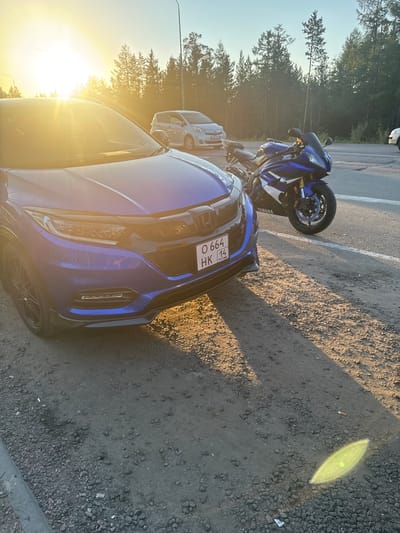 An eye-level, medium shot captures a blue Honda HRV and a blue Yamaha R6 motorcycle parked on a gravel shoulder beside a paved road. The setting is Serebryany Bor, Russia, during sunset. The sun, positioned in the upper left quadrant of the frame, casts a bright glare and long shadows across the scene, illuminating the dust and gravel. The car's front right quarter is in the foreground, angled towards the viewer, while the motorcycle is parked to its right, facing the road. A white vehicle is visible in the background to the left of the motorcycle, partially obscured by trees. The environment appears to be a roadside area with a dense forest bordering the right side of the road. The sky is clear and light blue, transitioning to a warmer tone near the horizon due to the setting sun.