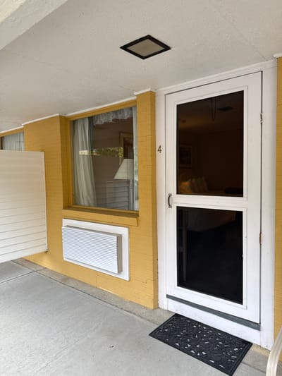 The image shows a ground-level view of an exterior entrance. The entrance features a white-framed door with two glass panes. Below the door is a window, also framed, and an adjacent white vent. A dark doormat sits to the right of the door. The walls are painted yellow and white. A small, framed black square is visible on the left wall. The ground is concrete. The address location within the city of Laconia in the United States of America cannot be determined from the visible elements of the image.