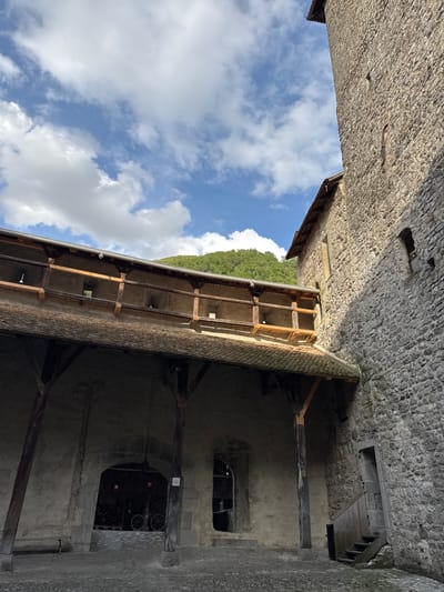 The image shows an upward view of a courtyard within Chillon Castle, located in Veytaux, Switzerland. The scene is framed by stone walls on both sides, built of rough-hewn blocks. On the right is the castle's interior, showing a structure built of stone with wooden beams supporting a shingled roof. A covered wooden walkway extends to the left, providing a covered exterior corridor. The sky is visible above with scattered white clouds and clear blue patches, and a green wooded hillside is visible in the background. The lighting is diffused, creating shadows within the castle structure and reflecting off the stone. The interior wall shows a window and a lower doorway with visible wagon wheels and a sign with an upward pointing arrow and "EXIT" in both english and french.