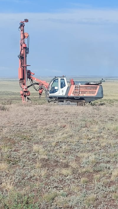 A red and white drilling rig is positioned in a dry, grassy field under a bright blue sky with some scattered clouds. The drilling rig is mounted on a tracked vehicle and has a large orange mast extending upwards. There appears to be a person in the cab of the rig. In the background, the landscape is flat and appears to be undeveloped, with some distant structures visible on the horizon. The overall impression is one of industrial activity in a remote, arid environment. The exact location in Saran, Kazakhstan is not determinable from the image itself.