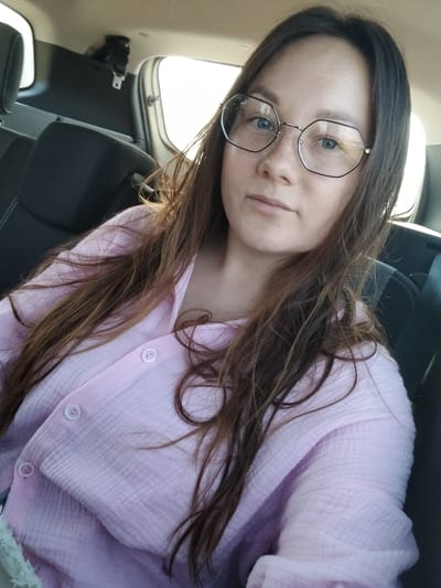 A young woman with long, brown hair and blue eyes, wearing glasses and a pink button-up shirt, is sitting in the back seat of a car. The car appears to be parked in a residential area, with houses and trees visible in the background. The woman is looking directly at the camera, and her expression is neutral. The overall scene is casual and relaxed, suggesting a candid moment.
